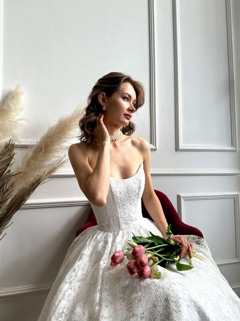 May include: A strapless white lace wedding dress with a full skirt. The dress is paired with a pearl necklace and earrings. A bouquet of pink and white tulips is held in the lap. The background features a white wall with decorative molding.