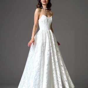 May include: A strapless white lace wedding dress with a fitted bodice and a full, flowing skirt. The dress is floor-length and features intricate floral lace detailing. The model is wearing a pearl necklace.