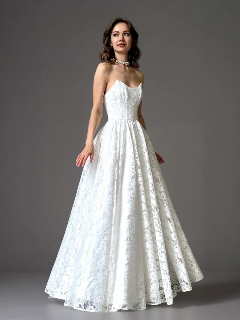 May include: A strapless white lace wedding dress with a fitted bodice and a full, flowing skirt. The dress is floor-length and features intricate floral lace detailing. The model is wearing a pearl necklace.