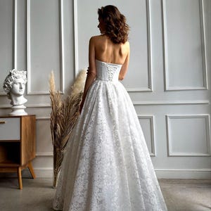 May include: A strapless white lace wedding dress with a full skirt and corset back. The dress is floor-length and features intricate floral lace detailing. The bride is facing away from the camera, showcasing the dress's design.