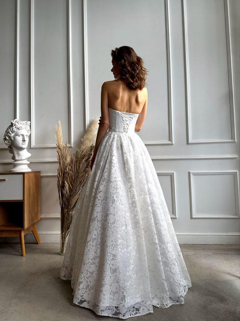 May include: A strapless white lace wedding dress with a full skirt and corset back. The dress is floor-length and features intricate floral lace detailing. The bride is facing away from the camera, showcasing the dress's design.