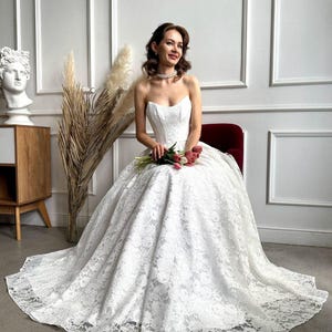 May include: A strapless white lace wedding dress with a full skirt and a long train. The bride is seated on a red chair and holding a bouquet of pink and red tulips. The dress features intricate floral lace detailing.