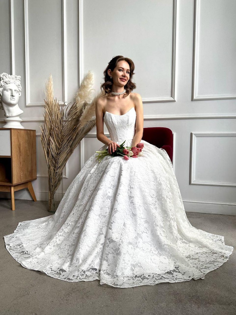 May include: A strapless white lace wedding dress with a full skirt and a long train. The bride is seated on a red chair and holding a bouquet of pink and red tulips. The dress features intricate floral lace detailing.