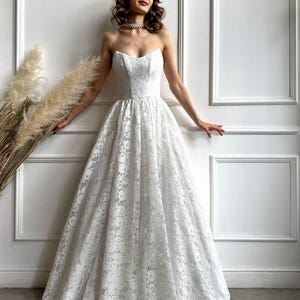 May include: A strapless white lace wedding dress with a sweetheart neckline and a full skirt. The dress is floor-length and features intricate floral lace detailing. The bride is wearing a pearl necklace.