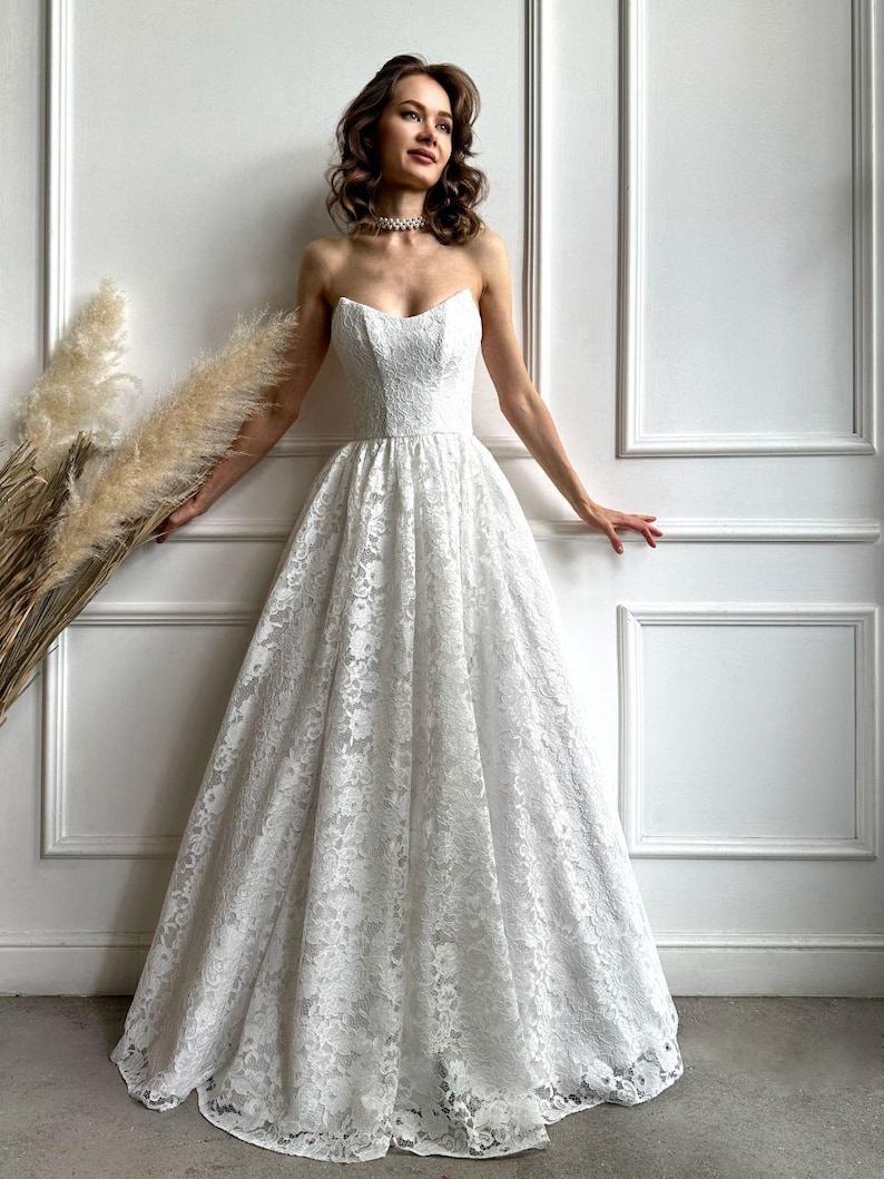 May include: A strapless white lace wedding dress with a sweetheart neckline and a full skirt. The dress is floor-length and features intricate floral lace detailing. The bride is wearing a pearl necklace.