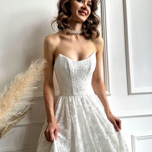 May include: A strapless white lace wedding dress with a sweetheart neckline and a full skirt. The dress is adorned with intricate floral lace patterns. A pearl necklace is worn around the neck.