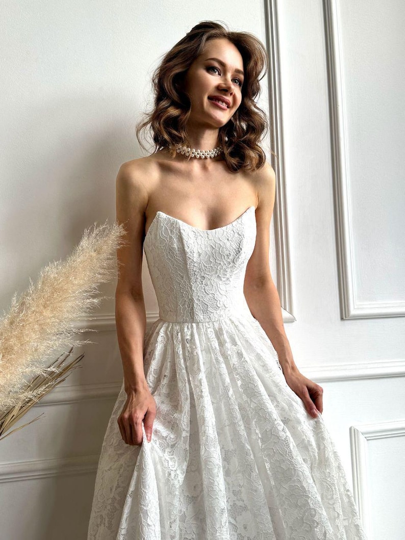 May include: A strapless white lace wedding dress with a sweetheart neckline and a full skirt. The dress is adorned with intricate floral lace patterns. A pearl necklace is worn around the neck.