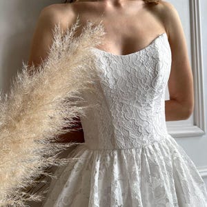 May include: A strapless white lace dress with a sweetheart neckline and a full skirt. The dress is adorned with intricate floral lace patterns. A pearl necklace is worn around the neck.