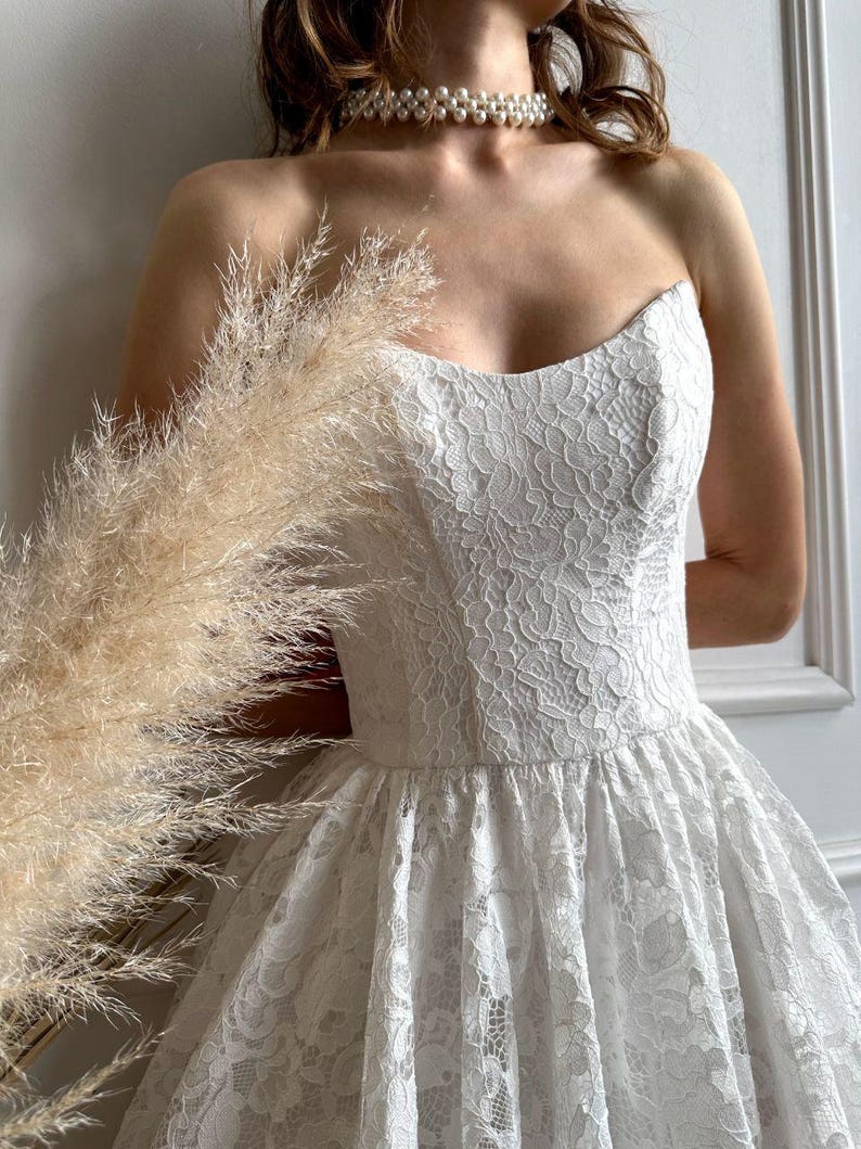 May include: A strapless white lace dress with a sweetheart neckline and a full skirt. The dress is adorned with intricate floral lace patterns. A pearl necklace is worn around the neck.