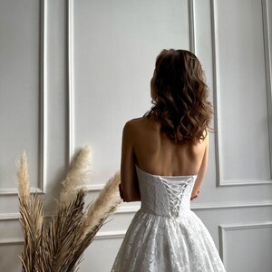 May include: A strapless white lace wedding dress with a corset back. The dress has a full skirt and is worn by a person with brown hair. The background is a white wall with vertical paneling.