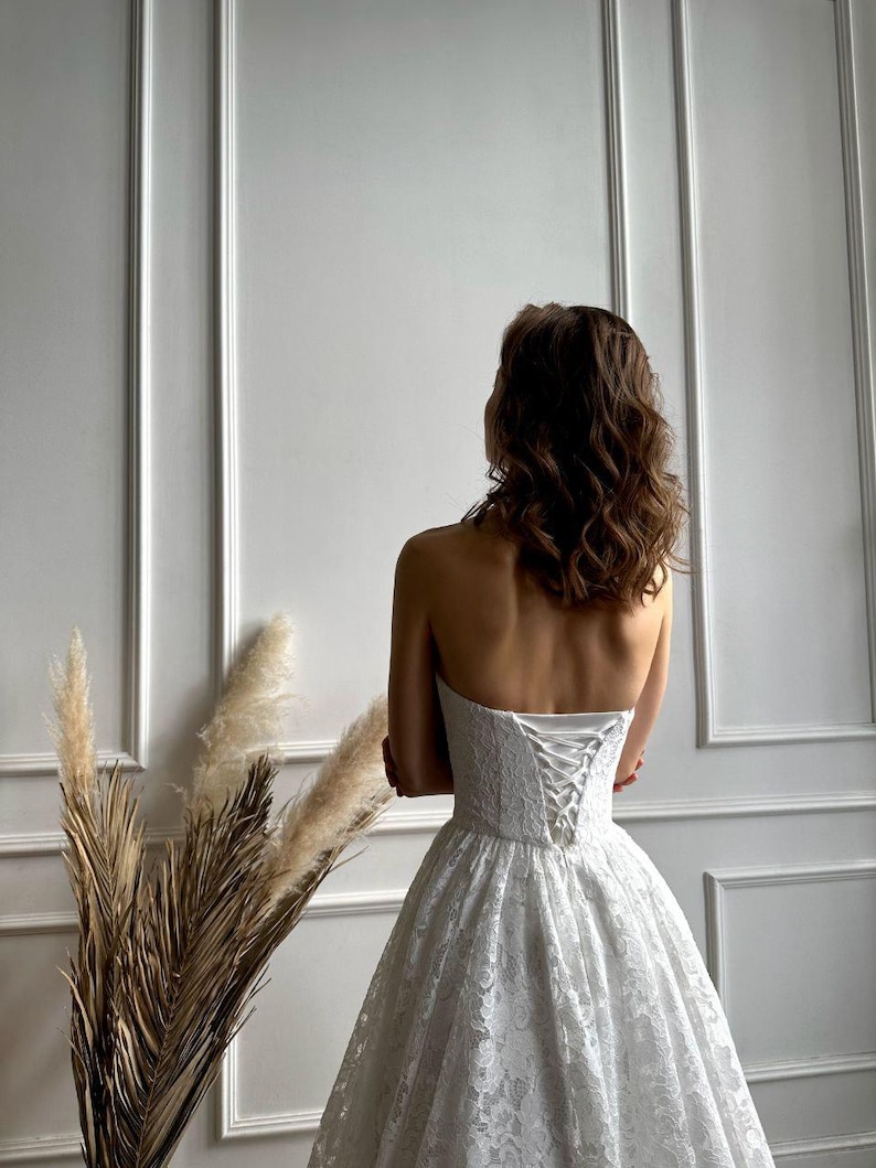 May include: A strapless white lace wedding dress with a corset back. The dress has a full skirt and is worn by a person with brown hair. The background is a white wall with vertical paneling.