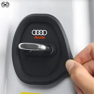 May include: A black silicone cover for a vehicle door latch, featuring the white Audi logo and the word Audi in orange text. The cover is shaped to fit around a metallic silver door latch mechanism, providing a protective layer for the car door.