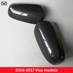 May include: A pair of side mirror covers with a black carbon fiber pattern, designed for 2014-2017 Vios models. The covers feature a glossy finish with a diagonal weave texture and smooth, rounded edges for a sleek automotive exterior look.