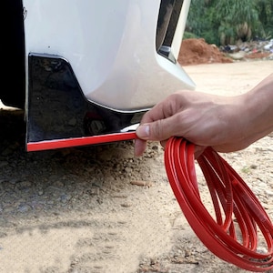 Könnte beinhalten: Ein flexibler, roter Klebestreifen, der an der Unterkante eines glänzend schwarzen Autostoßfänger-Bauteils angebracht wird. Der Streifen hat eine leuchtend rote Außenfläche und eine abziehbare Rückseite für die Fahrzeugdekoration.
