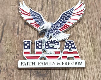 May include: A metal emblem featuring a bald eagle with wings spread, colored in red, white, and blue. Below the eagle, the letters USA are displayed in the same patriotic colors. A horizontal bar at the base bears the text FAITH, FAMILY & FREEDOM.