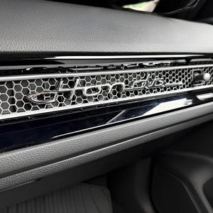 May include: A black dashboard interior featuring a silver honeycomb mesh vent pattern with the word Honda in a chrome script emblem centered across the horizontal trim piece, set against a textured black leather-like surface.