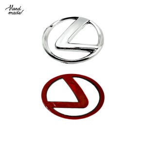 Pode incluir: Dois emblemas de carro Lexus. O emblema superior é prateado e cromado, com um "L" estilizado dentro de um círculo. O emblema inferior é vermelho com uma borda preta, também com o design "L". As palavras "Hand made" estão escritas em itálico no canto superior esquerdo.
