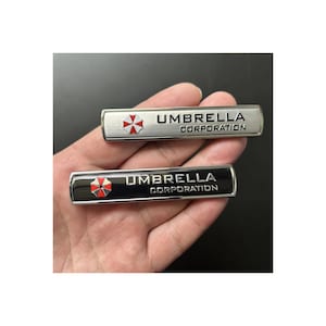 May include: Two rectangular metal badges with the text "UMBRELLA CORPORATION". The top badge is silver with a red and white logo, the bottom badge is black with the same logo. The words "hand made" are above the badges.