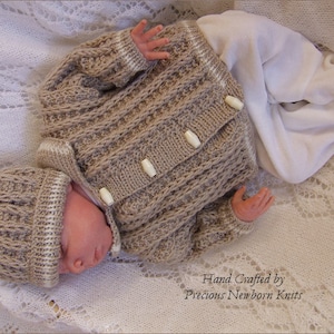 May include: A tan and cream knitted baby outfit, comprising a jacket with three white buttons and a matching hat. The jacket features a textured pattern, and the hat has a ribbed design. The text "Hand Crafted by Precious Newborn Knits" is visible.