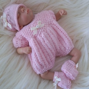 Puede incluir: Una muñeca bebé vestida con un conjunto de punto rosa claro, que incluye un vestido, un gorro y botines. El vestido y el gorro están adornados con lazos blancos. La muñeca está acostada sobre una superficie blanca y esponjosa.