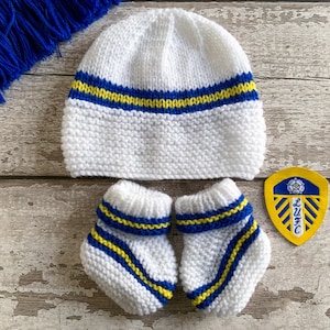 May include: A white knitted baby hat and booties set with blue and yellow stripes. A matching patch with a crest and the letters LUFC is also included. The set is displayed on a wooden surface.