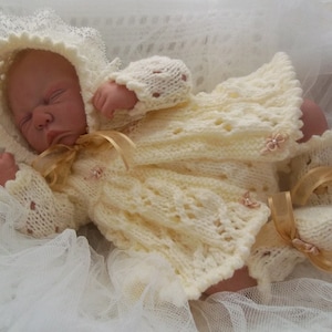 May include: A white crocheted baby outfit with a bonnet, dress, and booties. The outfit is decorated with small white flowers and gold ribbon.