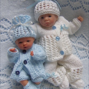 May include: Two baby dolls wearing hand-knitted outfits. One doll is in a light blue outfit with a matching hat and blue buttons. The other doll is in a white outfit with a matching hat, blue buttons, and a blue ribbon.