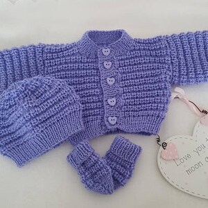 Puede incluir: Un conjunto de punto para beb&eacute; en color lila claro, que incluye un c&aacute;rdigan, un gorro y unas botitas. El c&aacute;rdigan tiene cierre de botones y botones en forma de coraz&oacute;n. El conjunto se muestra sobre una superficie blanca con un cartel en forma de coraz&oacute;n que dice "Love you to the moon and back".