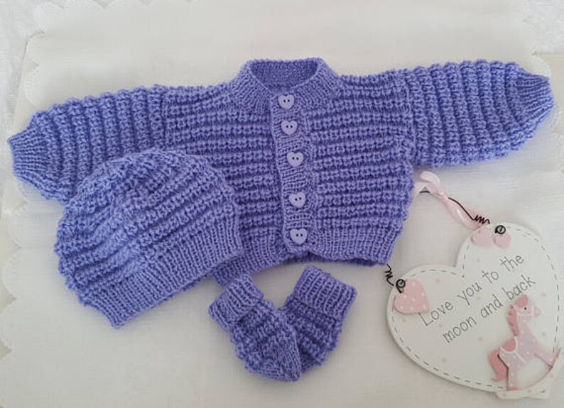Puede incluir: Un conjunto de punto para beb&eacute; en color lila claro, que incluye un c&aacute;rdigan, un gorro y unas botitas. El c&aacute;rdigan tiene cierre de botones y botones en forma de coraz&oacute;n. El conjunto se muestra sobre una superficie blanca con un cartel en forma de coraz&oacute;n que dice "Love you to the moon and back".