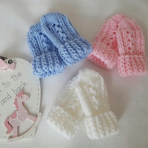 May include: Three pairs of knitted baby mittens in light blue, pink, and white. The mittens have a ribbed cuff and a textured pattern on the body. A decorative wooden sign with a pink rocking horse is in the background.