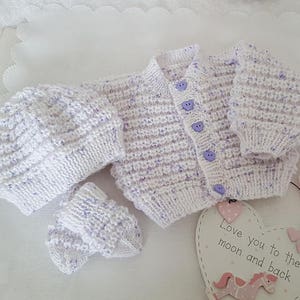 Puede incluir: Un conjunto de beb&eacute; de punto blanco con motas moradas. El conjunto incluye un gorro, un c&aacute;rdigan y botines. El c&aacute;rdigan tiene botones morados. El conjunto est&aacute; colocado sobre una superficie blanca con un letrero en forma de coraz&oacute;n que dice "Te quiero hasta la luna y de regreso".