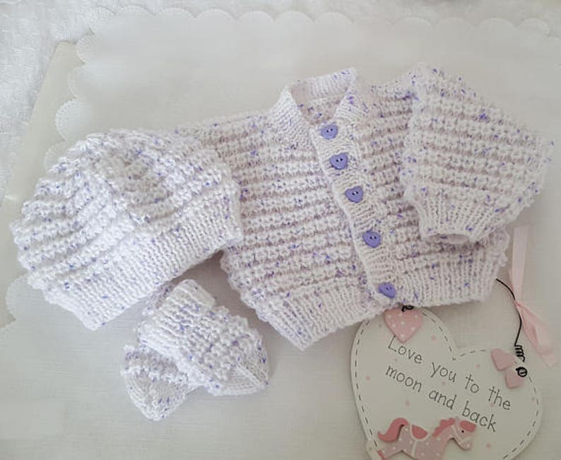 Puede incluir: Un conjunto de beb&eacute; de punto blanco con motas moradas. El conjunto incluye un gorro, un c&aacute;rdigan y botines. El c&aacute;rdigan tiene botones morados. El conjunto est&aacute; colocado sobre una superficie blanca con un letrero en forma de coraz&oacute;n que dice "Te quiero hasta la luna y de regreso".