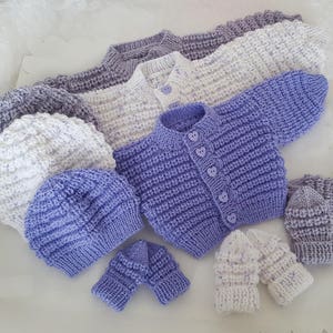 Puede incluir: Un conjunto de ropa de beb&eacute; de punto en tonos de morado y blanco, que incluye un c&aacute;rdigan, dos gorros y dos pares de guantes.