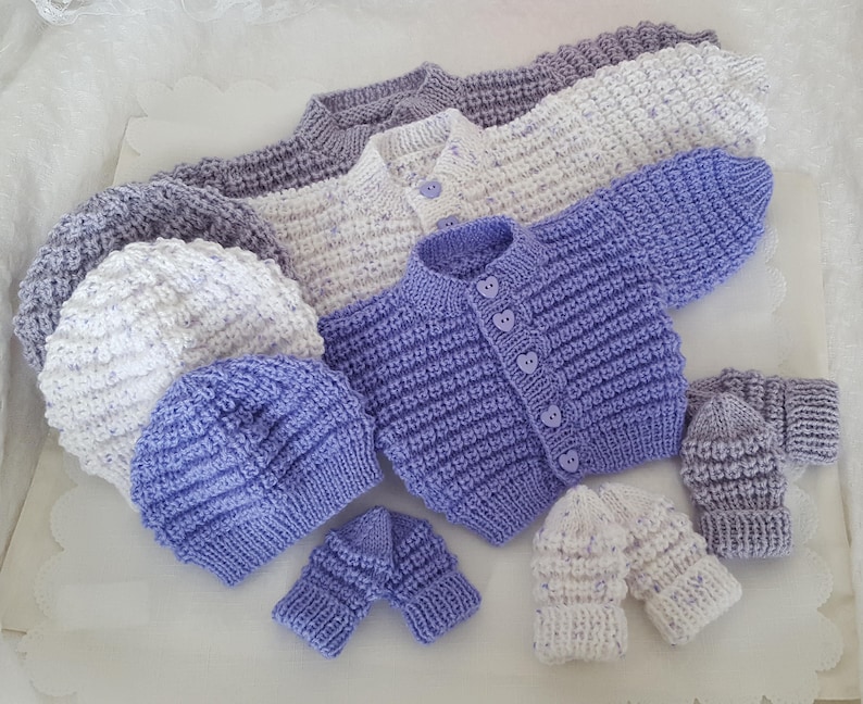Puede incluir: Un conjunto de ropa de beb&eacute; de punto en tonos de morado y blanco, que incluye un c&aacute;rdigan, dos gorros y dos pares de guantes.