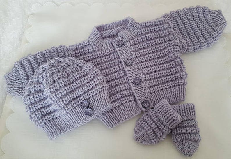 Puede incluir: Un conjunto de punto de beb&eacute; de color lavanda con gorro y botines a juego. El conjunto presenta un cierre de bot&oacute;n y un patr&oacute;n de punto texturizado.