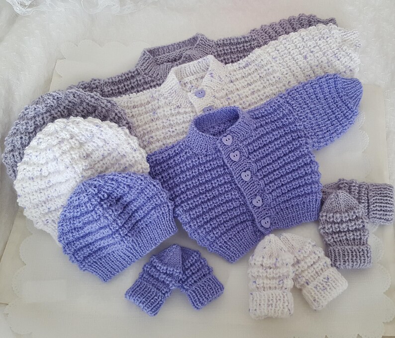Puede incluir: Un conjunto de ropa de beb&eacute; de punto en color morado y blanco, que incluye un c&aacute;rdigan, dos gorros y dos pares de guantes.