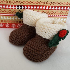 May include: A pair of hand-knitted booties featuring a brown textured base and a thick, cream-colored garter stitch cuff. Each bootie is decorated with a small, crocheted green holly leaf and a single red berry accent on the outer side.