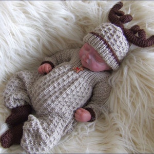 May include: A baby wearing a brown and beige knitted reindeer onesie and hat with brown antlers. The baby is lying on a white fluffy surface.