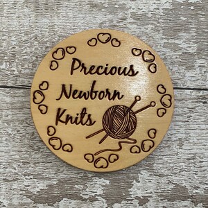 Puede incluir: Un letrero redondo de madera con el texto "Precious Newborn Knits" y una imagen estilizada de un ovillo de lana y agujas de tejer. El letrero est&aacute; rodeado por un borde de peque&ntilde;os corazones.