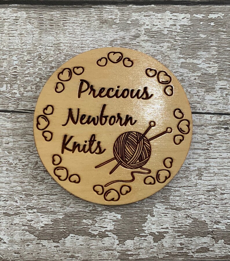 Puede incluir: Un letrero redondo de madera con el texto "Precious Newborn Knits" y una imagen estilizada de un ovillo de lana y agujas de tejer. El letrero est&aacute; rodeado por un borde de peque&ntilde;os corazones.