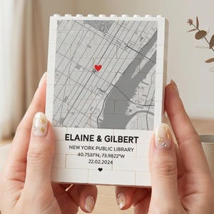 May include: A white block with a map of New York City, featuring a red heart marking a location. The block has the names "Elaine & Gilbert", location details, and a date printed on it. The block is held by a person.