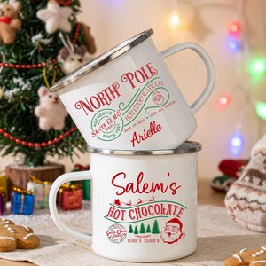 Puede incluir: Dos tazas de esmalte blanco con bordes plateados. La taza superior presenta texto en rojo y verde que dice North Pole Hot Chocolate y el nombre Arielle. La taza inferior presenta texto en rojo y verde que dice Salem's Hot Chocolate.