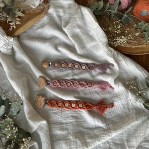 Könnte beinhalten: Drei Baby-Schnullerhalter aus Makramee in Lila-, Rosa- und Orangetönen. Jeder Halter hat Holzperlen mit einem Namen und einen Holzclip in Tierform. Die Halter werden auf einem weißen Stoff präsentiert.
