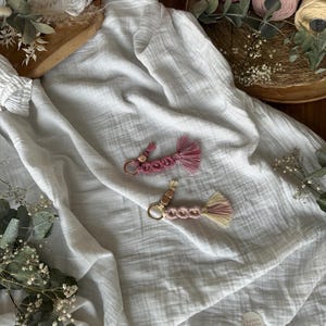 Könnte beinhalten: Zwei handgefertigte Makramee-Schlüsselanhänger mit rosa und beigen Quasten und Holzperlen. Die Schlüsselanhänger befinden sich auf einem weißen Stoffhintergrund mit Trockenblumen und Garnrollen im Hintergrund. Die Schlüsselanhänger haben goldfarbene Schlüsselringe.