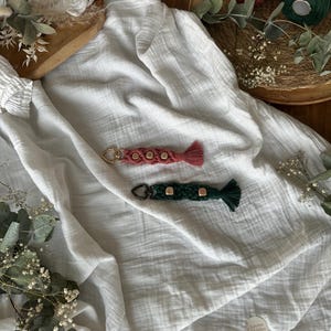 Op de afbeelding: Twee macramé sleutelhangers, één koraal en één donkergroen, elk met houten letterkralen die een naam spellen. De sleutelhangers hebben een kwast aan het uiteinde en liggen op een witte stof. De sleutelhangers zijn handgemaakt.