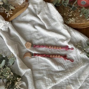 May include: Two pink pacifier clips with wooden beads spelling out names. One clip has a flower-shaped wooden piece, the other a crescent moon. The clips rest on a white, textured fabric with floral accents.