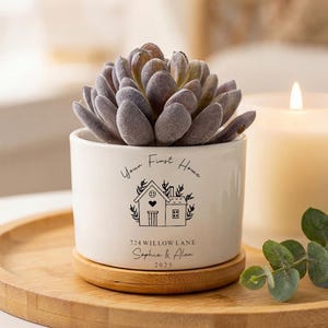 May include: A white ceramic planter with a faux succulent, resting on a wooden coaster. The planter has a black illustration of a house and the text "Your First Home", "724 Willow Lane", "Sophia & Alex", and "2025". A lit candle and greenery are in the background.