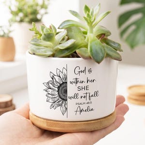 May include: A white ceramic planter with a sunflower design and the text "God is within her. She will not fall. Psalm 46:5 Amelia." The planter holds a succulent plant and sits on a bamboo saucer. Other plants are in the background.