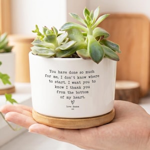 May include: A white ceramic cylindrical planter with a bamboo saucer base containing a green succulent plant. The front of the planter features black printed text that reads: You have done so much for me, I don't know where to start. I want you to know I thank you from the bottom of my heart. Love Emma xx.