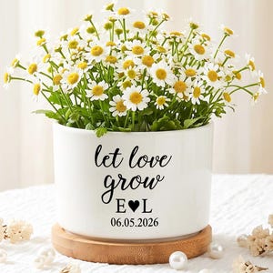May include: A white ceramic planter with the text "let love grow" and initials, filled with daisies. The planter sits on a wooden coaster, with pearls and flowers scattered around.